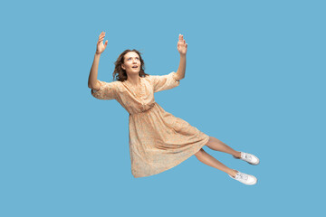 Hovering in air. Full length beautiful pensive girl in yellow dress levitating flying in mid-air, looking up with dreamy relaxed expression. indoor studio shot isolated on blue background - obrazy, fototapety, plakaty