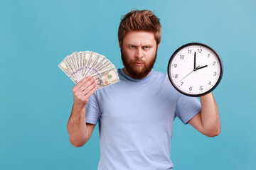 Portrait of angry handsome bearded man holding big fan of dollar banknotes and wall clock, time is...