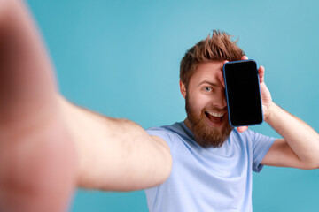 Portrait of positive optimistic bearded man blogger making selfie, point of view photo, covering eye with mobile phone, smiling happily. Indoor studio shot isolated on blue background.