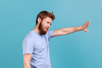 Side view of strict bearded man showing stop sign, standing with frowning face and bossy expression, meaning caution to avoid danger or mistake. Indoor studio shot isolated on blue background.