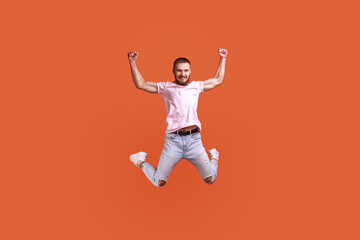 Fototapeta na wymiar Portrait of happy attractive bearded man jumping in air raised arms, looking at camera, copy space for ad, wearing pink T-shirt and jeans. Indoor studio shot isolated on orange background.