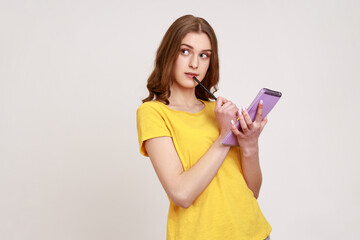 Beautiful thoughtful teen girl in yellow casual style T-shirt holding notebook in hand, writing...