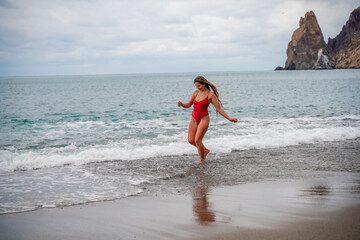 A beautiful and sexy brunette in a red swimsuit on a pebble beach, Running along the shore in the foam of the waves