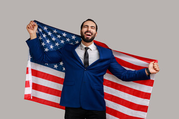 Excited bearded man holding USA flag and looking at camera with rejoice look, celebrating national holiday, wearing official style suit. Indoor studio shot isolated on gray background.
