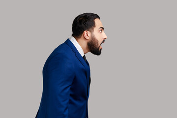 Side view of surprised astonished bearded man, standing with open mouth, being impressed of shocking news, wearing official style suit. Indoor studio shot isolated on gray background.