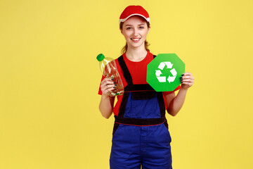 Fototapeta na wymiar Smiling positive worker woman volunteer standing with empty plastic bottle and green recycling sign, wearing overalls and red cap. Indoor studio shot isolated on yellow background.