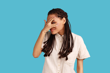 Portrait of interested woman with dreadlocks covering eyes with hand, peeking through fingers with happy curious expression, wearing white shirt. Indoor studio shot isolated on blue background.