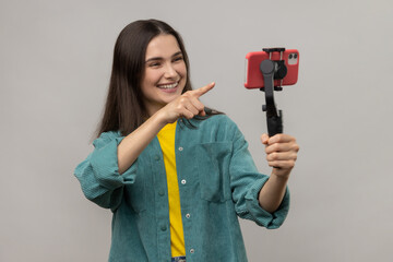 Portrait of smiling woman pointing to cell phone camera with smile, streaming, talking with followers, uses steadicam, wearing casual style jacket. Indoor studio shot isolated on gray background.