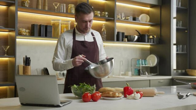 Young man in apron prepares dinner following video. Chef cooks dish with vegetables mixing sauce in bowl and watching recipe in laptop at table in kitchen
