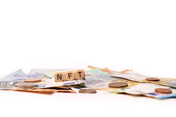 NFT spelled with wooden letters wooden cube on a plain white background with banknotes and coins, concept image