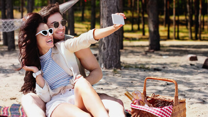 Happy couple in sunglasses taking selfie on smartphone near basket on beach