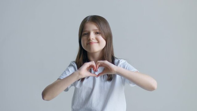 A blonde teenager girl in a white T-shirt smiles and folds her hands like a heart against a white wall