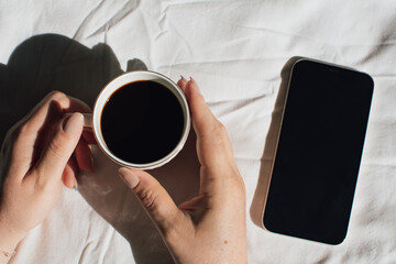 A woman working from bed, holding a coffee and with a cell phone