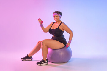 Size plus fitness model, athletic trainer instructor woman in home gym sit on fitball isolated on pink background. Workout sport concept - obrazy, fototapety, plakaty