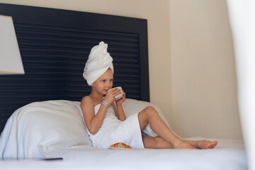 A little girl in a white towel drinks morning coffee in bed at home