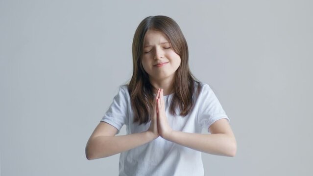 A blonde teenager girl in a white T-shirt prays for a wish in front of a white wall