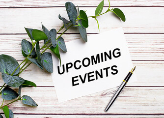 On a light wooden table, there is a eucalyptus branch, a fountain pen and a sheet of paper with the text UPCOMING EVENTS