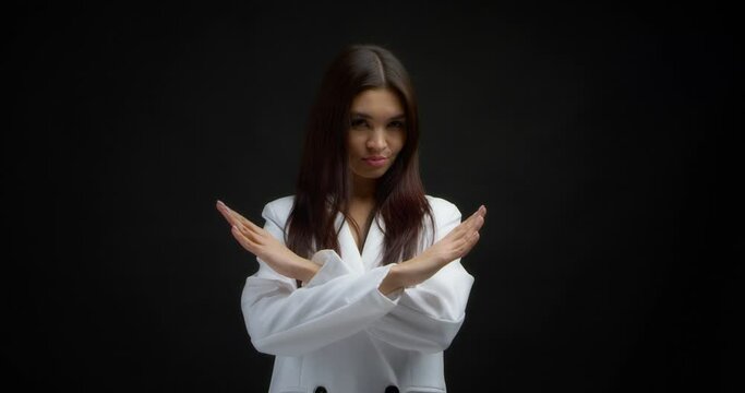 Angry young woman holds her crossed arms in an x gesture and looks at the camera. The stop sign, concept of negation. A strict serious businesswoman in a white jacket shows no on a black background