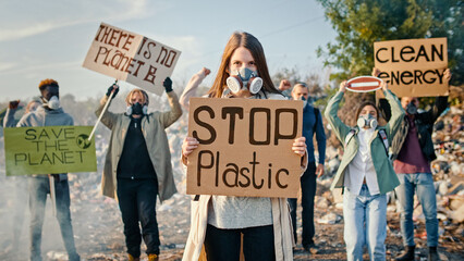 Portrait of Attractive Young Woman Activist Holding a Poster Calling to Stop Plastic. In Background Fighting People Protesting Against Garbage Pollution Staying at Dump in City Outskirts.