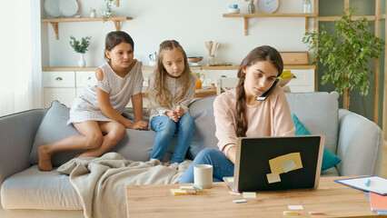 A Busy Mother Working Remotely With Her Children's at Home. The Daughters are Sits on the Couch, While Their Mother Working With a Laptop at the Table and Doing Business Conversation on the Phone.