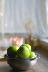 Bowl of green apples, open book, reading glasses, lit candle and flowers on the table. Selective focus.