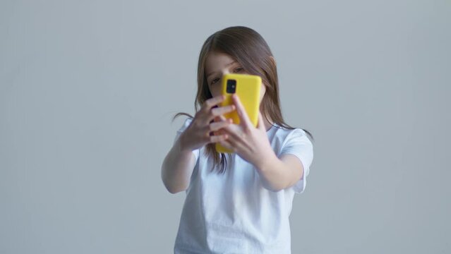 A blonde teenager girl in a white T-shirt takes a selfie in the phone against the background of a white wall