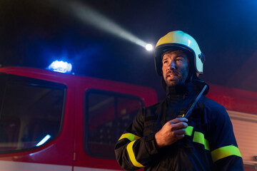 Low angle view of m firefighter talking to walkie talkie with fire truck in background at night.