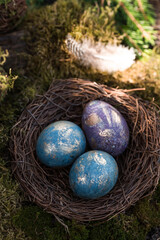 Happy Easter 2022. A postcard with a copy of the place for the text. Purple, blue and golden eggs in a nest on a natural forest background. Top view.