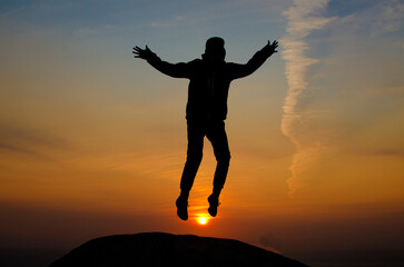 Young man jumping over a rock looking straight. Nature and beauty concept. Orange sundown. Man silhouette at sunset
