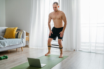 Fototapeta na wymiar Fit sporty healthy man on a gym mat using rubber resistance exercise band doing legs stretching exercises, watching online training class on a laptop. Healthy people and self-motivation concept.