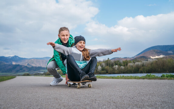 Laughing kids have fun using a skateboard. Brother pushing his sister sitting on a skateboard and making wings with arms. Happy outdoor childhood without an electronic devices concept image.