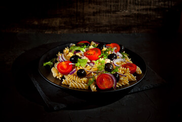 Salad with tomatoes, peppers, tuna olives and pasta. Fresh salad in a black plate on a dark background.