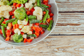 A mixture of frozen vegetables in a white bowl. Preparation for a dish on an old shabby table. Sliced vegetables on a light board background. Copy space and free space for text next to food.
