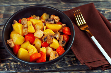 Boiled potatoes with meat in a black bowl. Potatoes with mushrooms and carrots. Boiled mushrooms and chicken with potatoes. Food on a black wooden table.