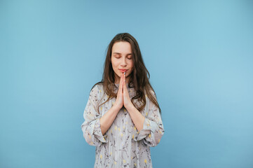 Beautiful woman in a dress stands on a blue background and prays with closed eyes and a cute smile.