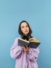 Beautiful girl in a sweatshirt and headphones reads a book on a blue background with a serious face. Vertical