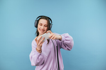 Positive female gamer in headset on head plays mobile online games on smartphone with smile on face isolated on blue background © bodnarphoto