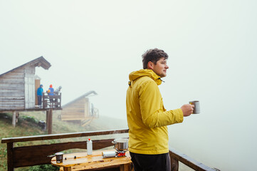 Male hiker in a yellow jacket stands on the balcony of a house in the mountains with a cup of hot drink in his hands on a background of foggy views.