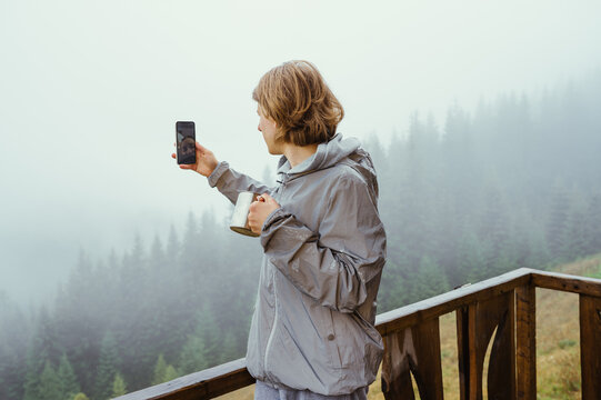 A male tourist in a jacket stands on the balcony of a dacha in the mountains with a cup in his hands and takes photos of beautiful misty landscapes.