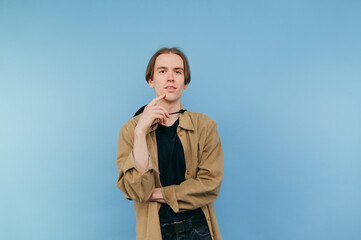 Handsome young man in a shirt stands on a blue background and looks at the camera with a pensive face and smiles.