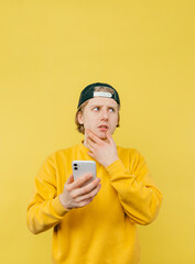 Portrait of a pensive guy in a sweatshirt and cap on a yellow background with a smartphone in his hand scratches his beard and looks away. Vertical