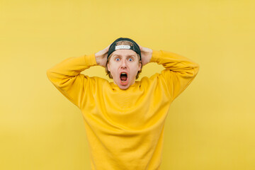 Portrait of a shocked guy in a cap and sweatshirt, holding hands behind his head and shouting in...