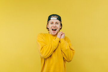 Portrait of joyful guy in cap and yellow sweatshirt standing happy on yellow background and looking at camera with open mouth and happy face.