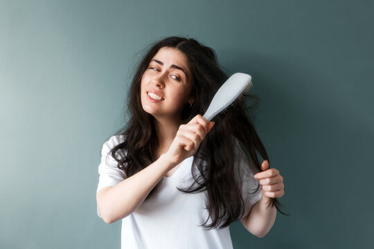 Portrait of a young sad woman trying to comb her long dark tangled hair with hairbrush. Blue background. The concept of unruly hair care