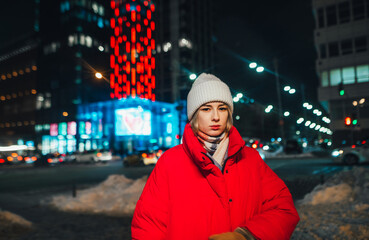 Beautiful woman in a red jacket and hat stands at night on the street on a background of snow-covered city and looks at the camera with a serious face.