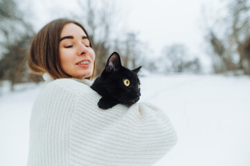 Positive woman in a white sweater with a black cat in her arms stands on the street in winter, holding a pet in her arms and smiling.
