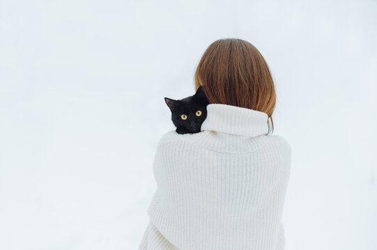 Back of woman in white light with black cat on shoulders, pet looking at camera.