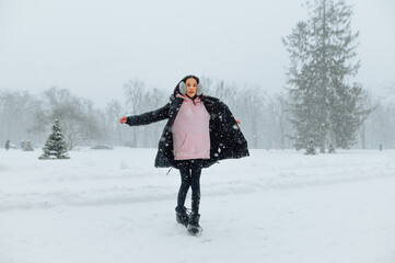 Happy hispanic woman in warm clothes having fun in the park during a heavy snowfall with a smile on her face, full length portrait.