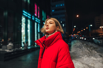 Fototapeta premium Portrait of a beautiful woman in a red jacket on the street at night, looking aside against the backdrop of the city evening landscape