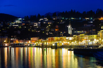View of embankment in Ascona, canton of Ticino, Switzerland. Houses along waterfront, turned on city lights in evening.